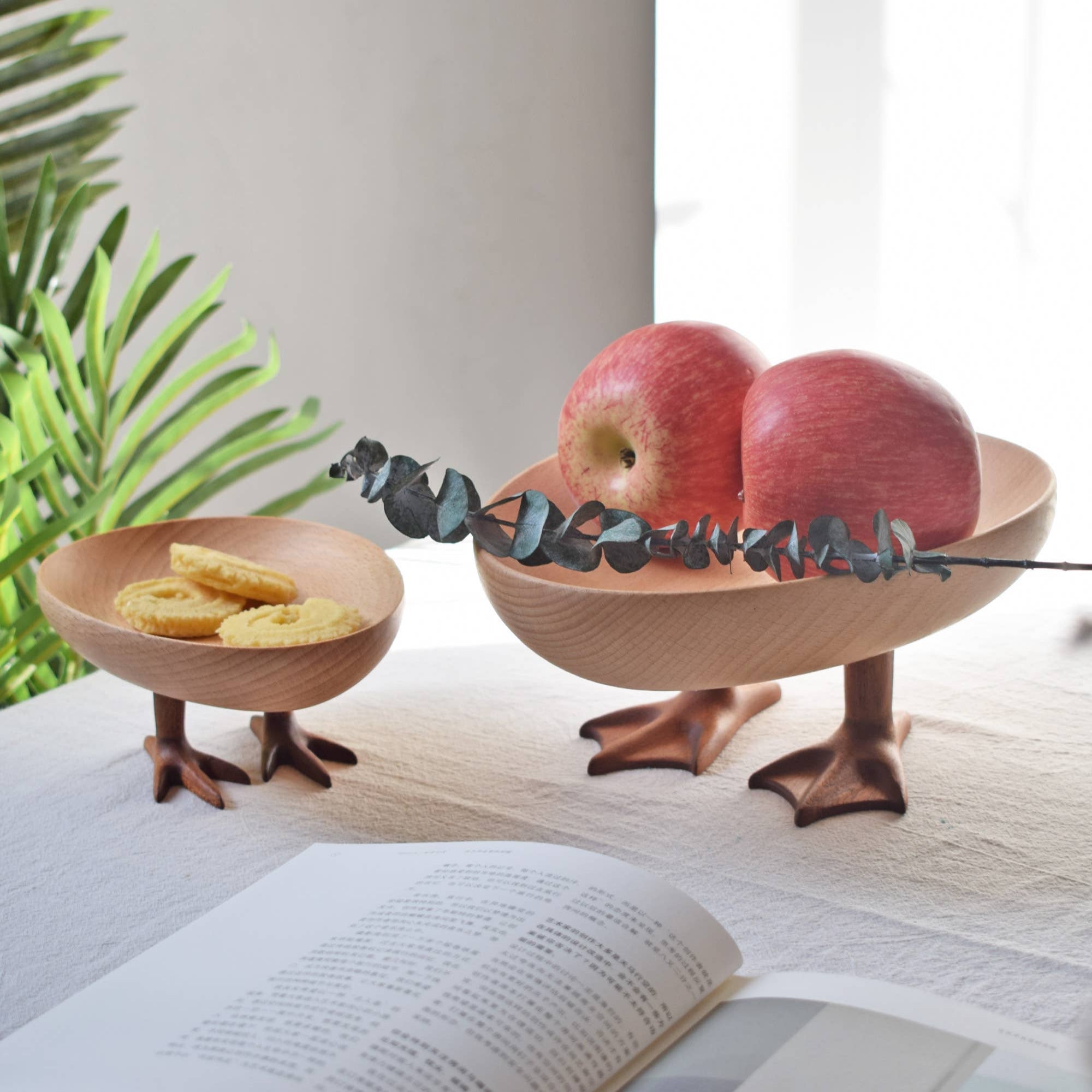 Two wooden bird-shaped bowls with apples and cookies on a surface next to an open book.