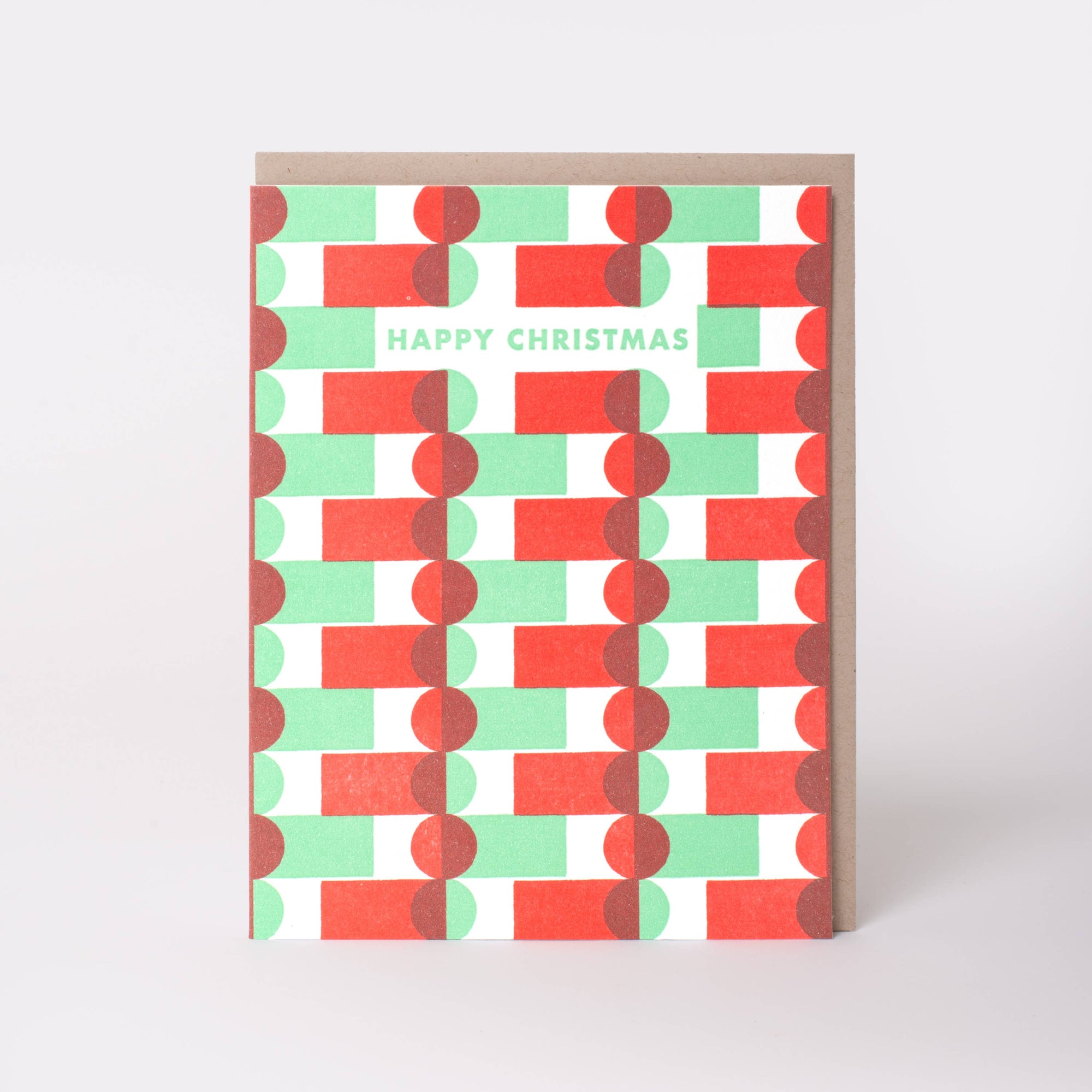 A Christmas greeting card with a geometric pattern of red, mint green, and white overlapping rectangles, featuring the text 'HAPPY CHRISTMAS' in mint green. The card is shown standing upright with a brown envelope behind it on a light gray background.