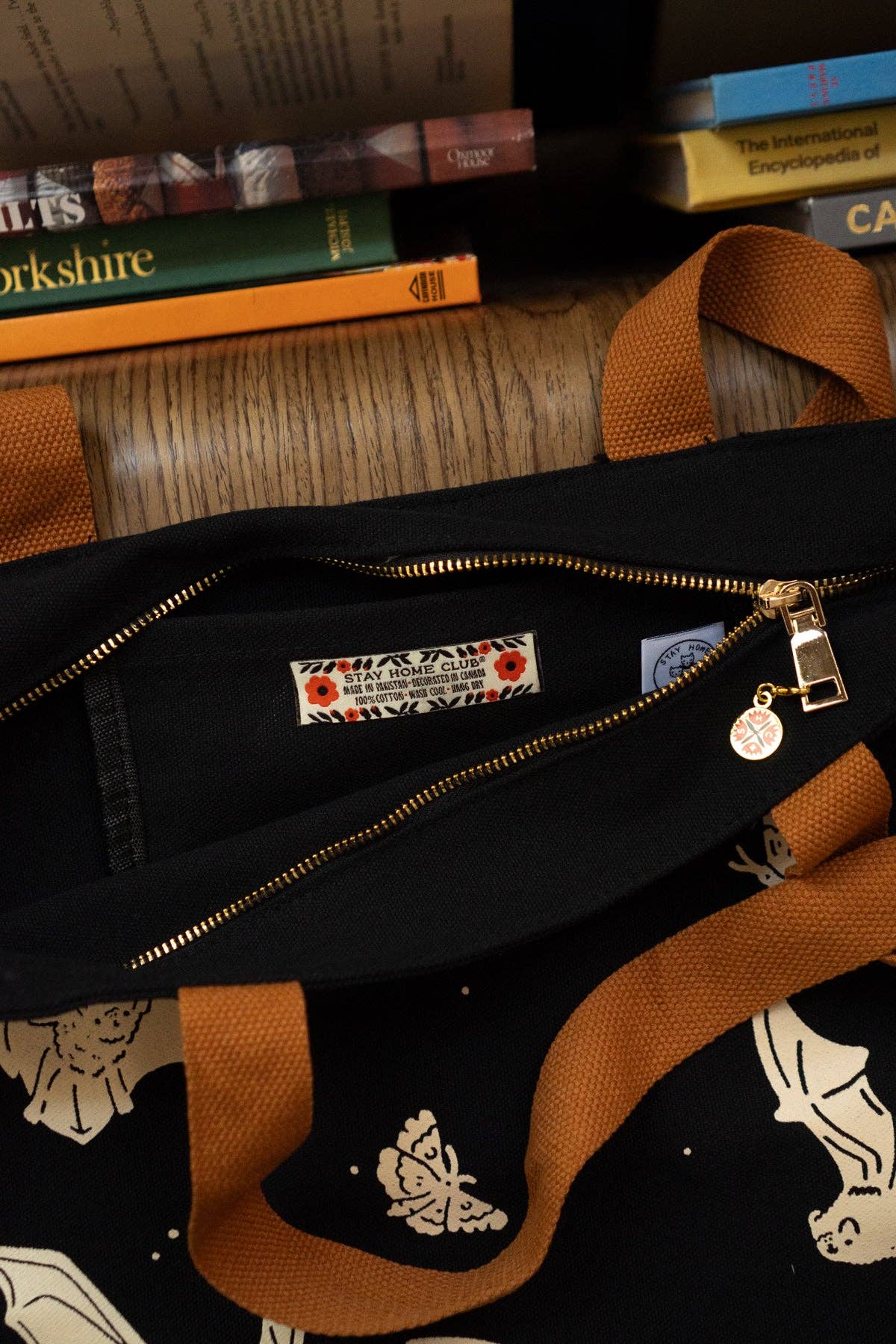 A close-up of a black canvas tote bag with brown canvas handles, gold zipper, and cream butterfly print, shown with a visible interior label and zipper pull on a wooden surface with books in the background.