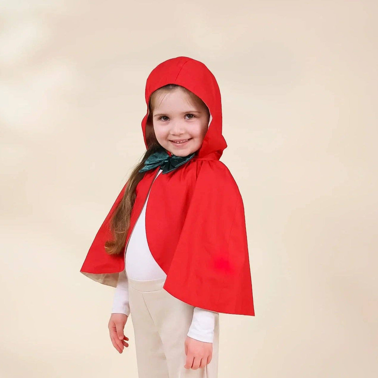 A young girl modeling a red hooded cape costume over a white outfit, standing against a plain beige studio background.