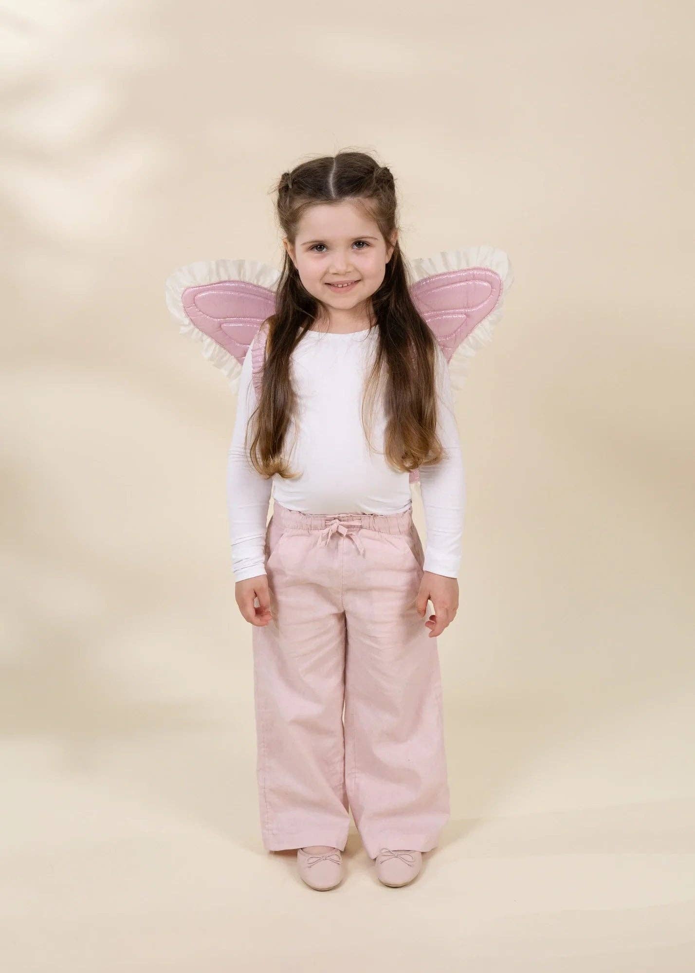 A young girl with long brown hair stands against a warm beige studio background wearing a white long-sleeve top, light pink wide-leg pants, and pink angel wings with white fringed trim.