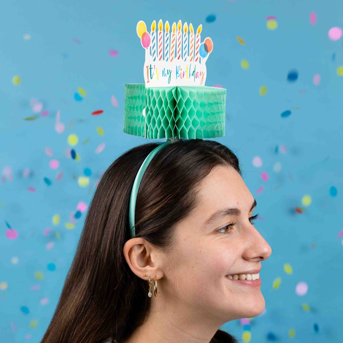 Person wearing a birthday cake headband with candles against a blue background with confetti.