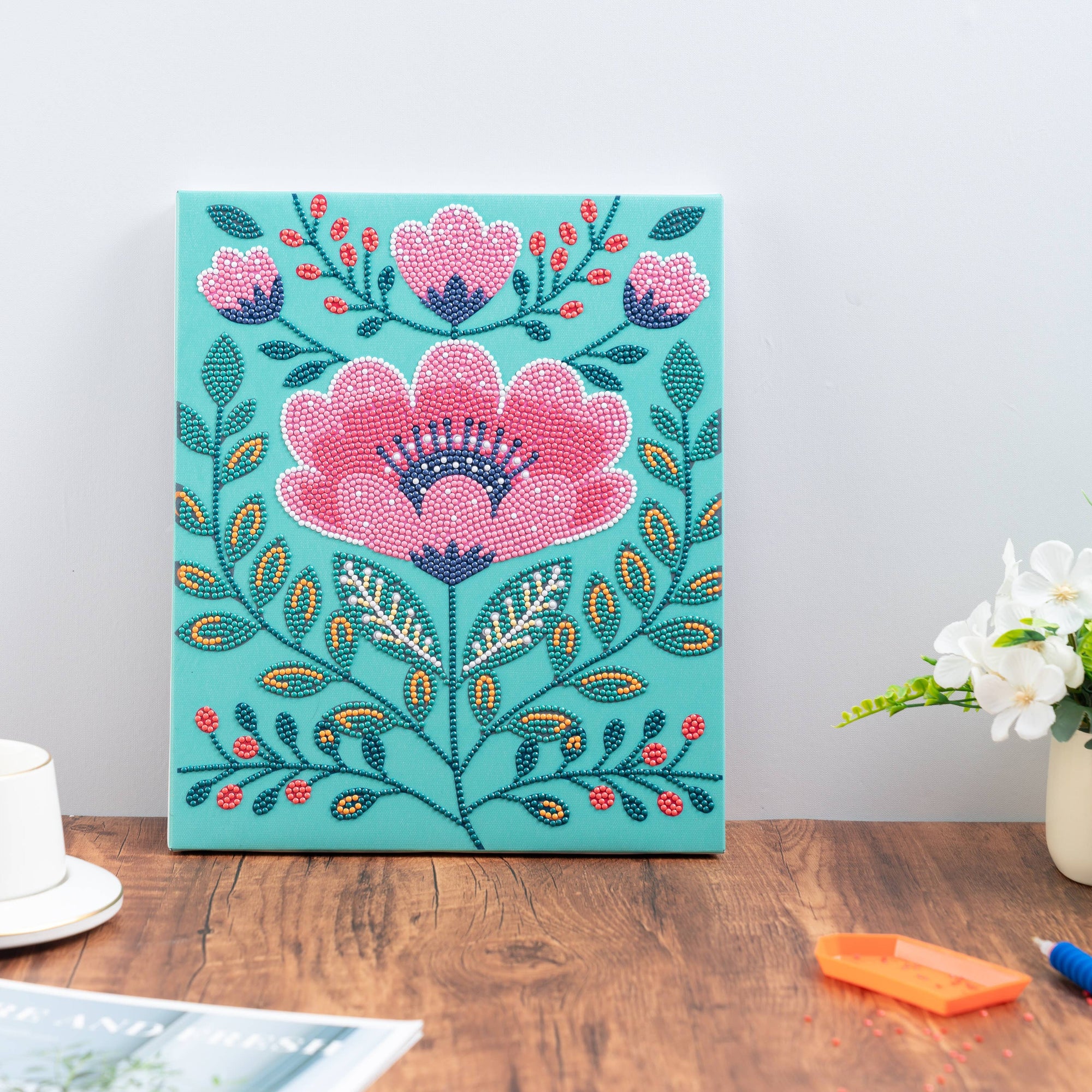 Decorative floral painting on a teal background with a cup and plant on a wooden table.