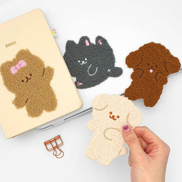 Four cute plush animal-shaped desk accessories in tan, black, brown, and cream are displayed on a white surface, including a notebook cover, a laptop stand, a binder clip, and a hand holding one cream dog accessory.