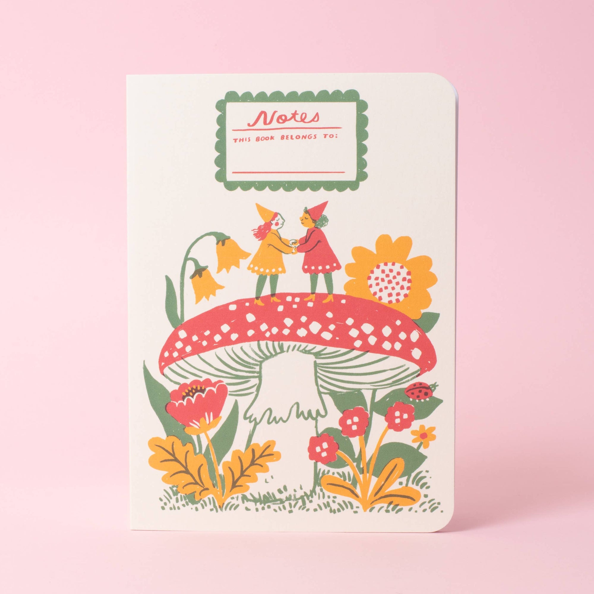 A white hardcover notebook with a whimsical folk-art illustration of two children on a red mushroom, surrounded by yellow flowers, set against a solid pink background.