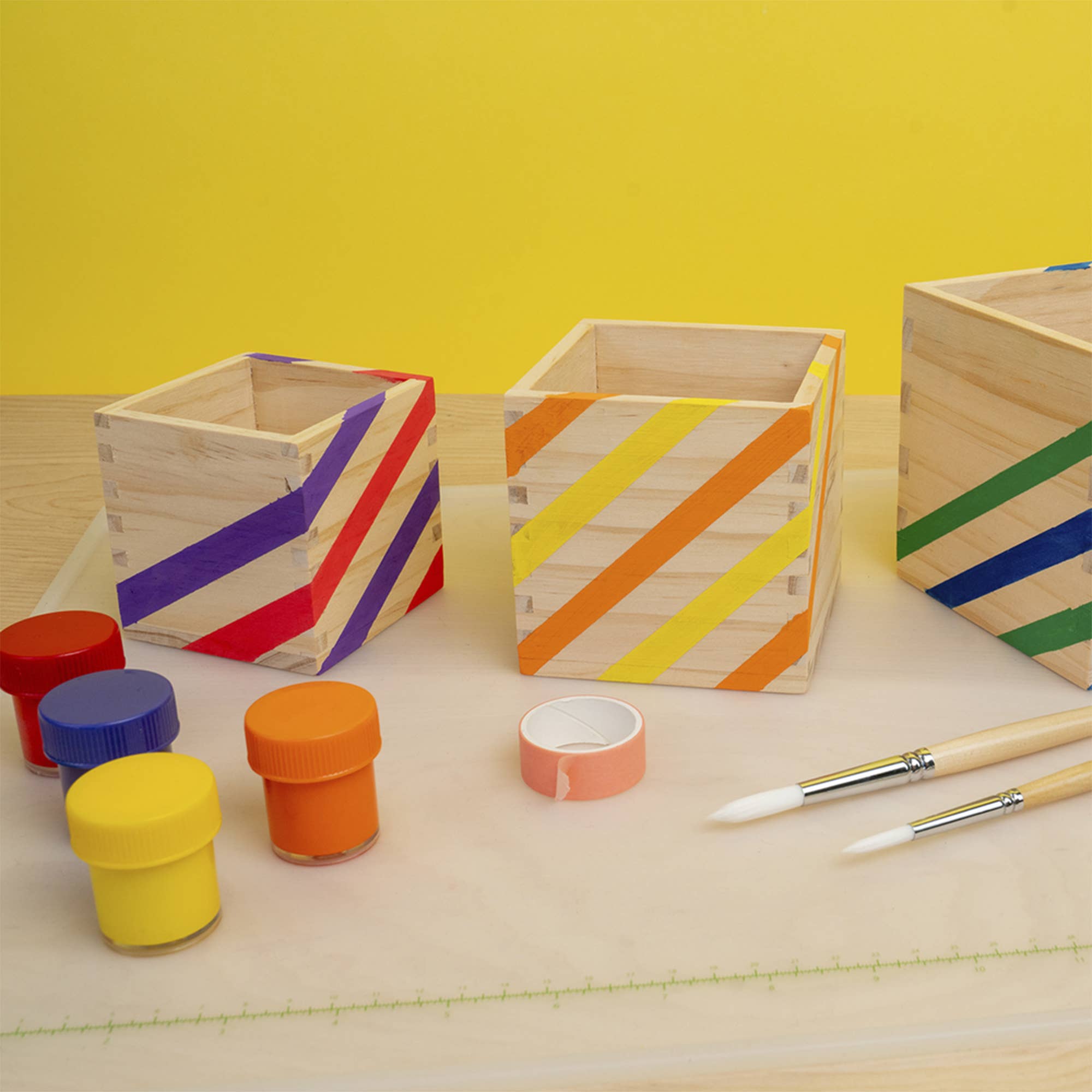 A children's craft kit featuring three wooden boxes decorated with colorful diagonal stripes (purple, red, yellow, orange, green, blue), four small jars of paint in yellow, orange, blue, and red, a roll of pink tape, and two paintbrushes on a light wood surface against a solid yellow background.