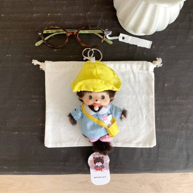 Small plush toy with a yellow raincoat on a white pouch with sunglasses and a white container in the background.