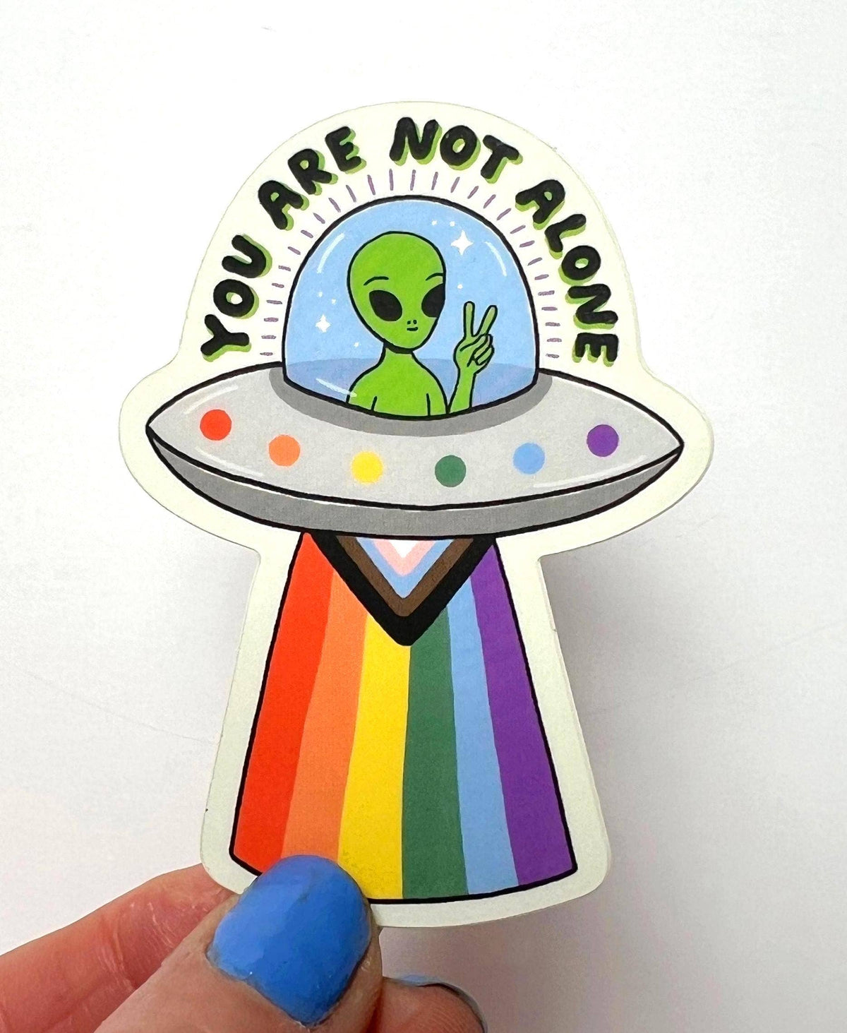 A die-cut sticker featuring a green alien in a UFO with the text 'YOU ARE NOT ALONE' above it, held by a hand with blue nail polish against a plain white background.