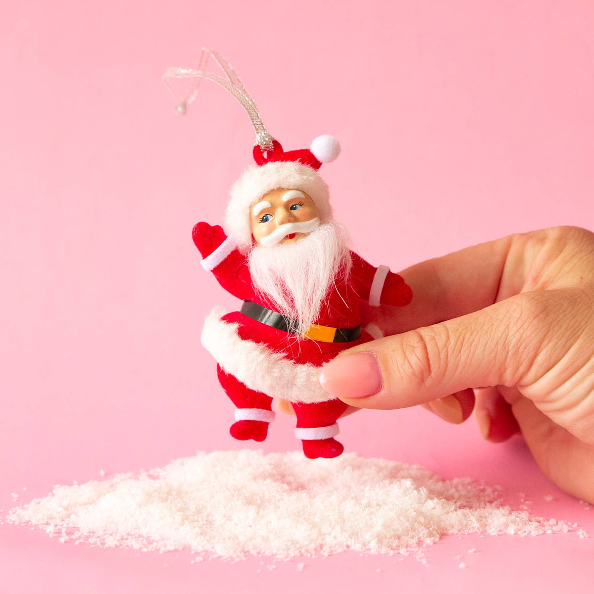 A hand holds a small Santa Claus hanging ornament above a pile of white faux snow against a solid pink background.