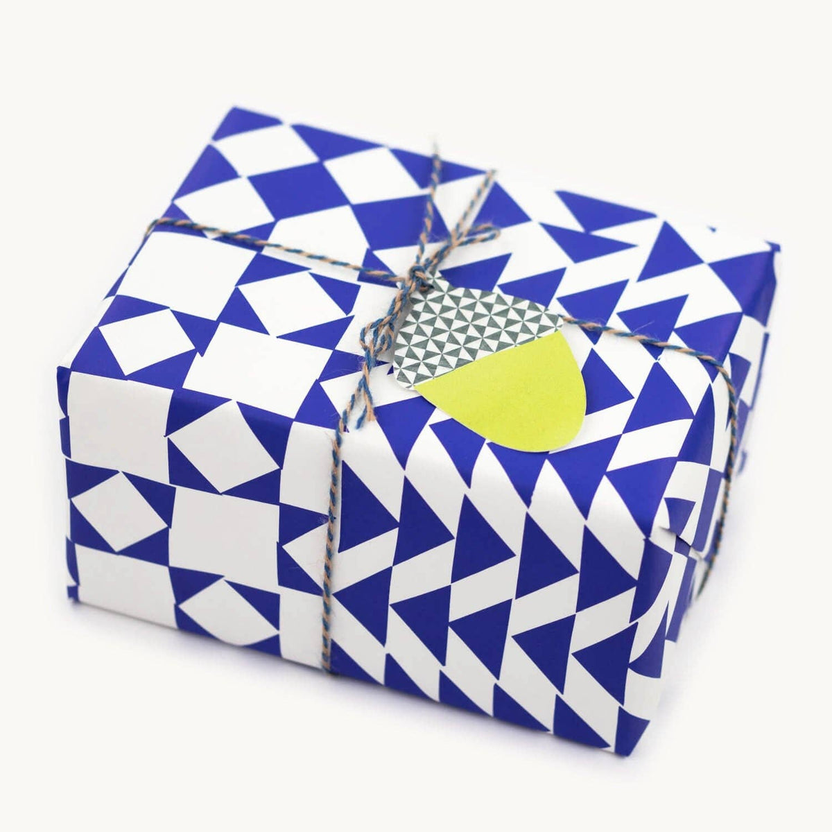 A square gift box wrapped in blue and white geometric patterned paper, tied with twine and a small paper tag, on a light off-white background.