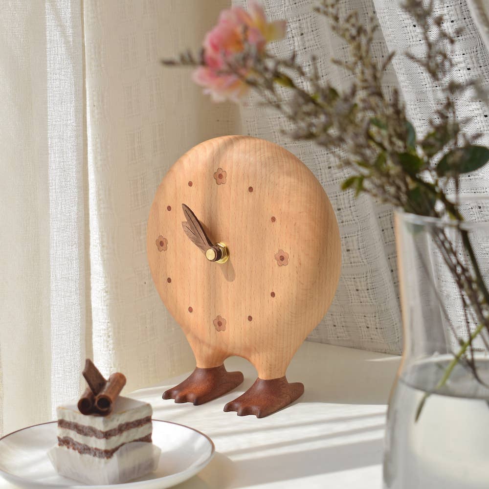 Wooden bird-shaped clock on a surface with a plate of cake and flowers in the background.