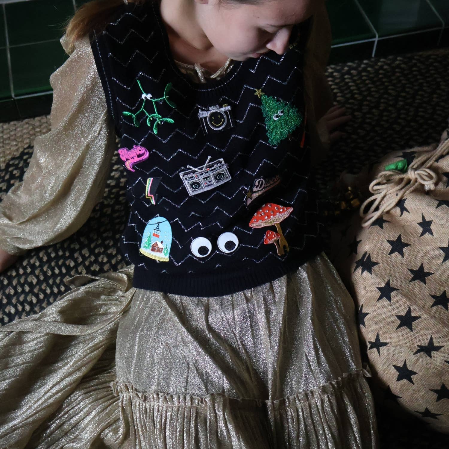 Child wearing a dark sweater with colorful patches sitting on a patterned surface.