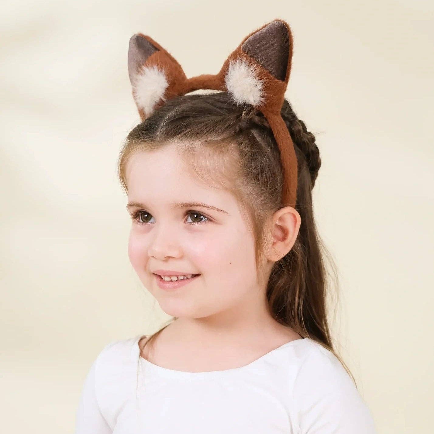 A young girl with brown hair in a braid wearing a brown faux fur fox ear headband, posing against a soft beige studio background.
