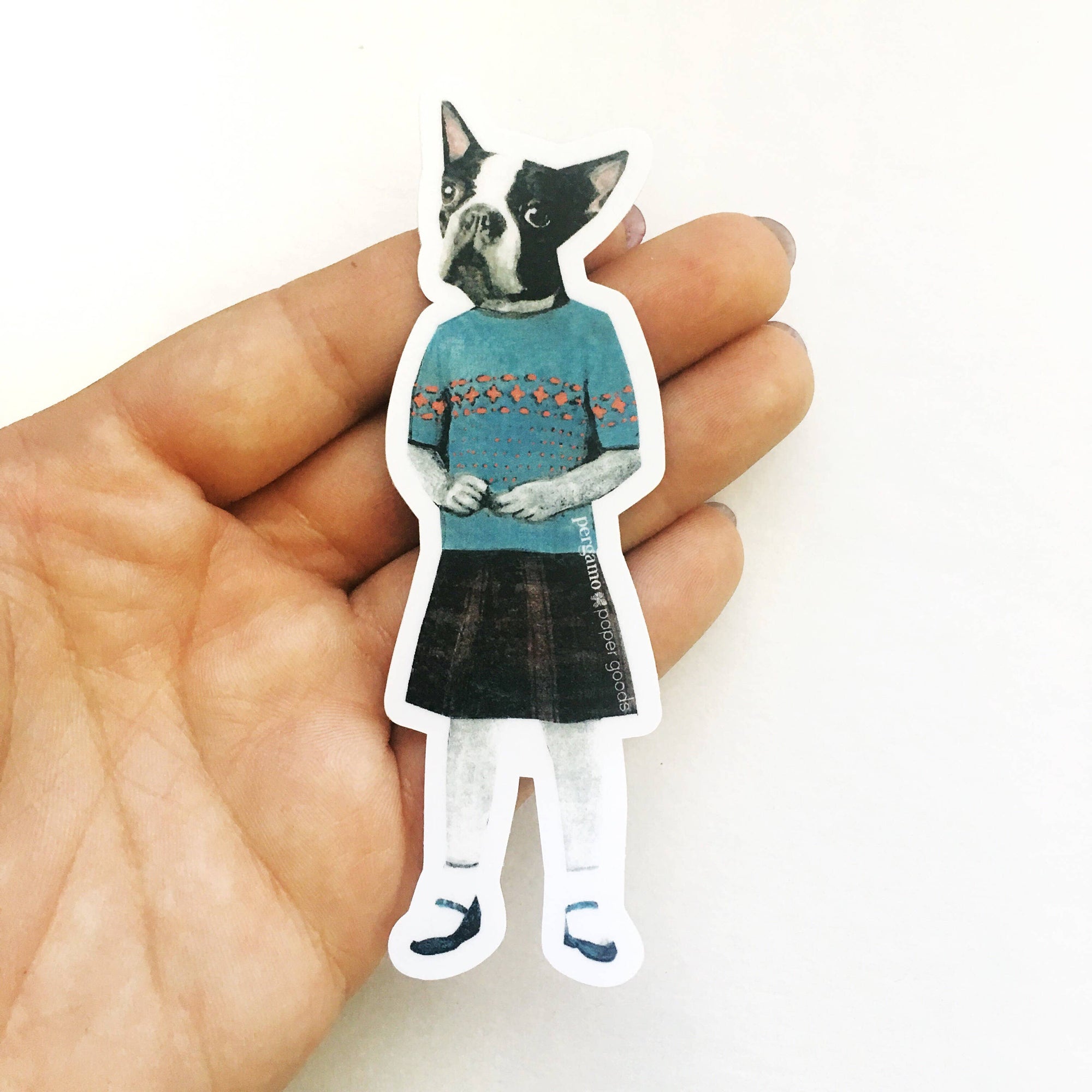 Hand holding a sticker of a dog wearing a sweater and skirt on a white background