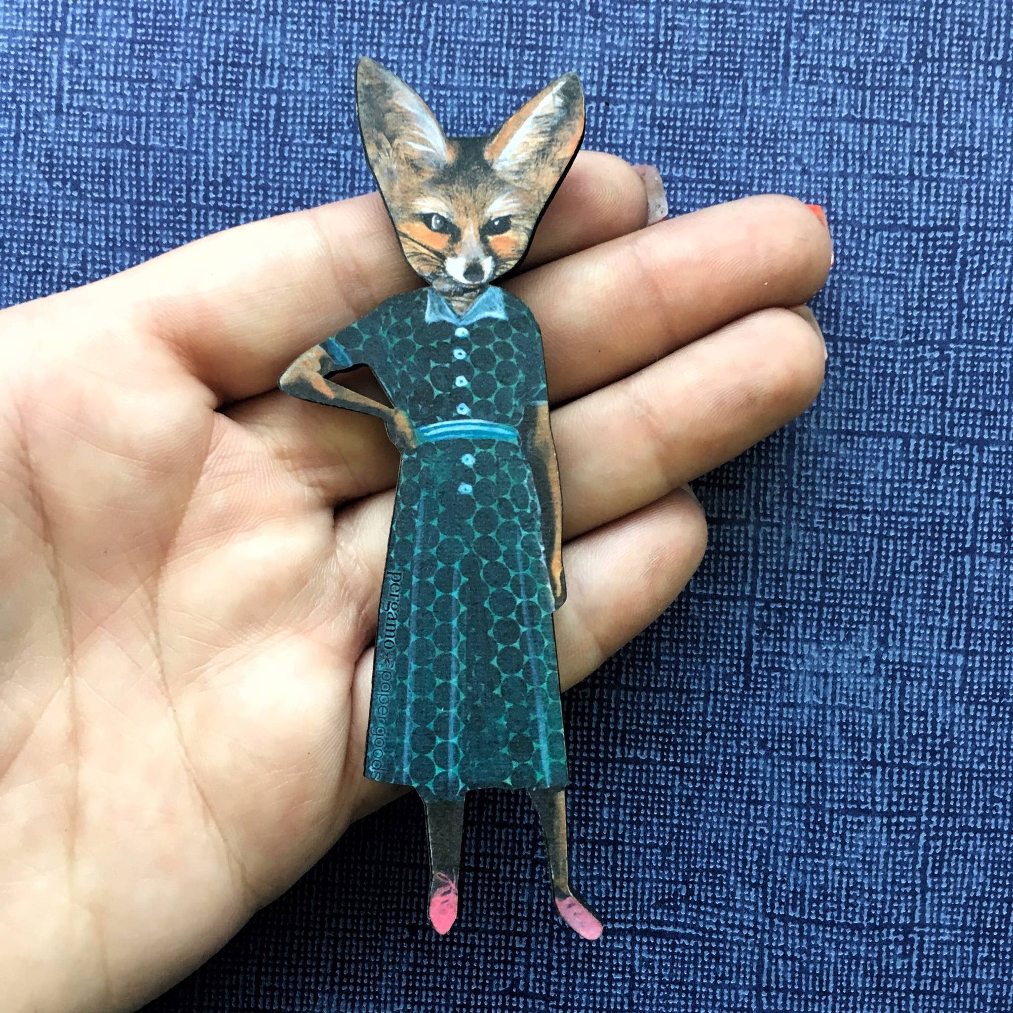 Hand holding a paper figurine of a fox in a dress against a blue textured background