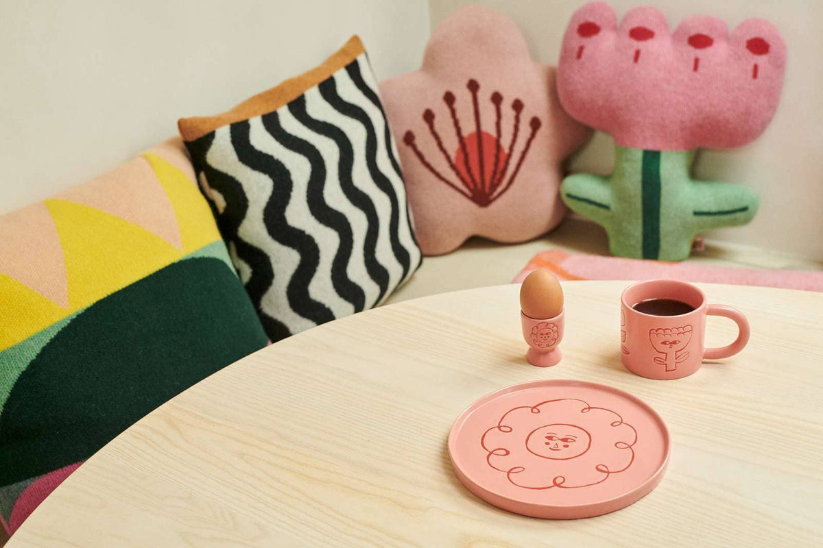 A cozy tabletop scene with a pink ceramic plate and matching mug featuring a simple line-drawn face and flower motif. A small egg cup with an egg and a dark coffee mug sit beside the plate on a light wood table.