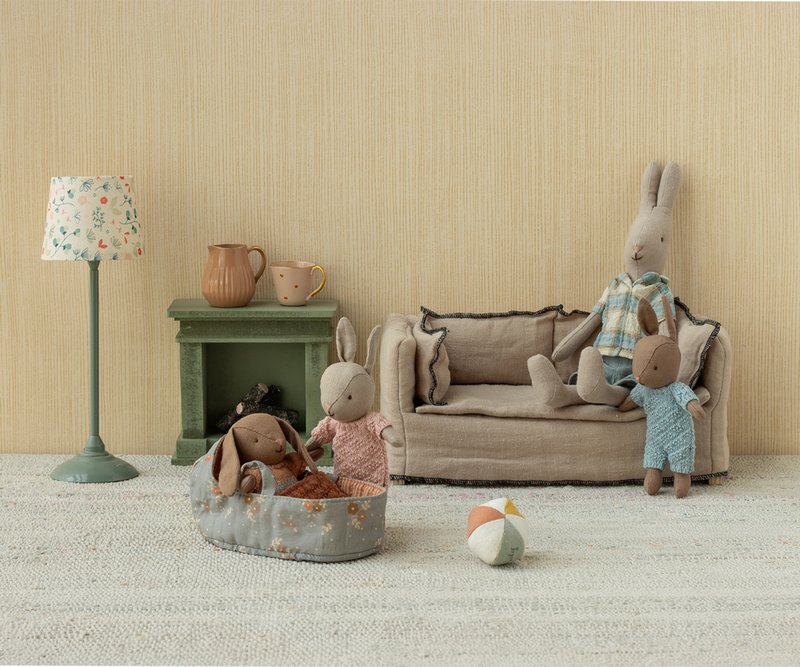 Toy rabbits and a rabbit in a plaid outfit in a room with a lamp, fireplace, and sofa.