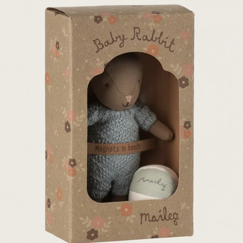A baby rabbit plush toy in a knitted blue outfit, displayed inside its brown kraft paper packaging box with floral accents. The box features a die-cut window showing the rabbit and a small ball.