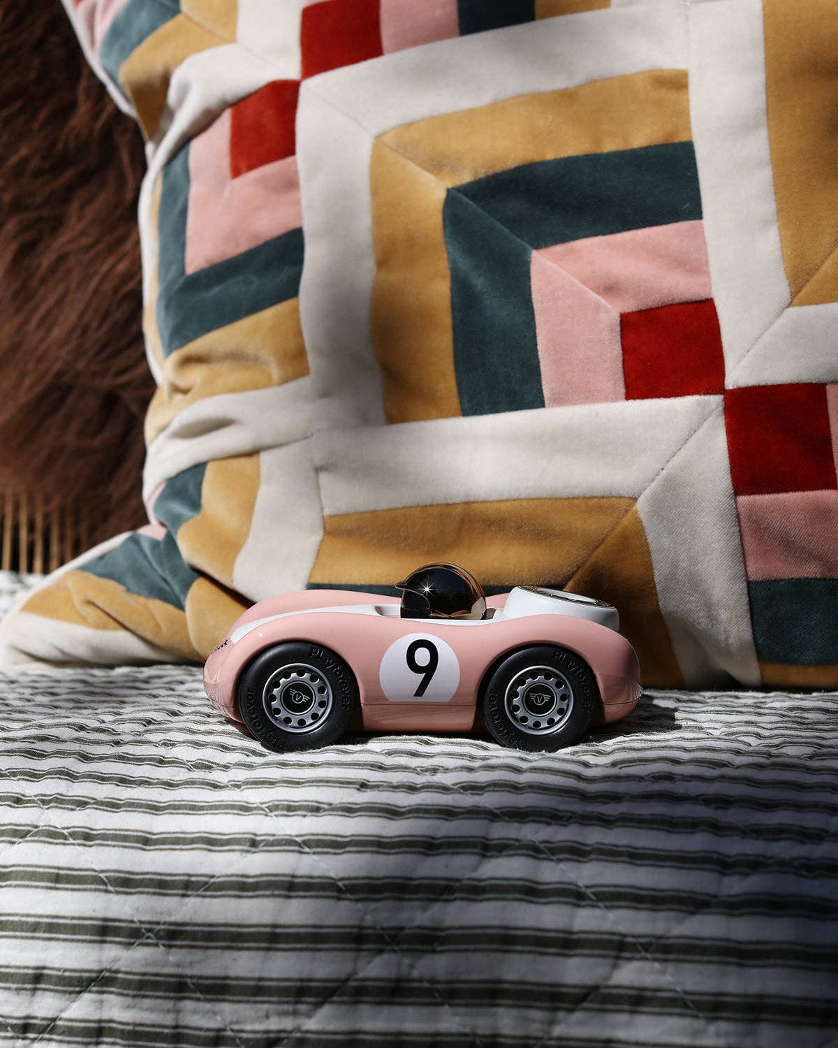 Pink toy car with number 9 on a striped surface, next to a colorful quilt.