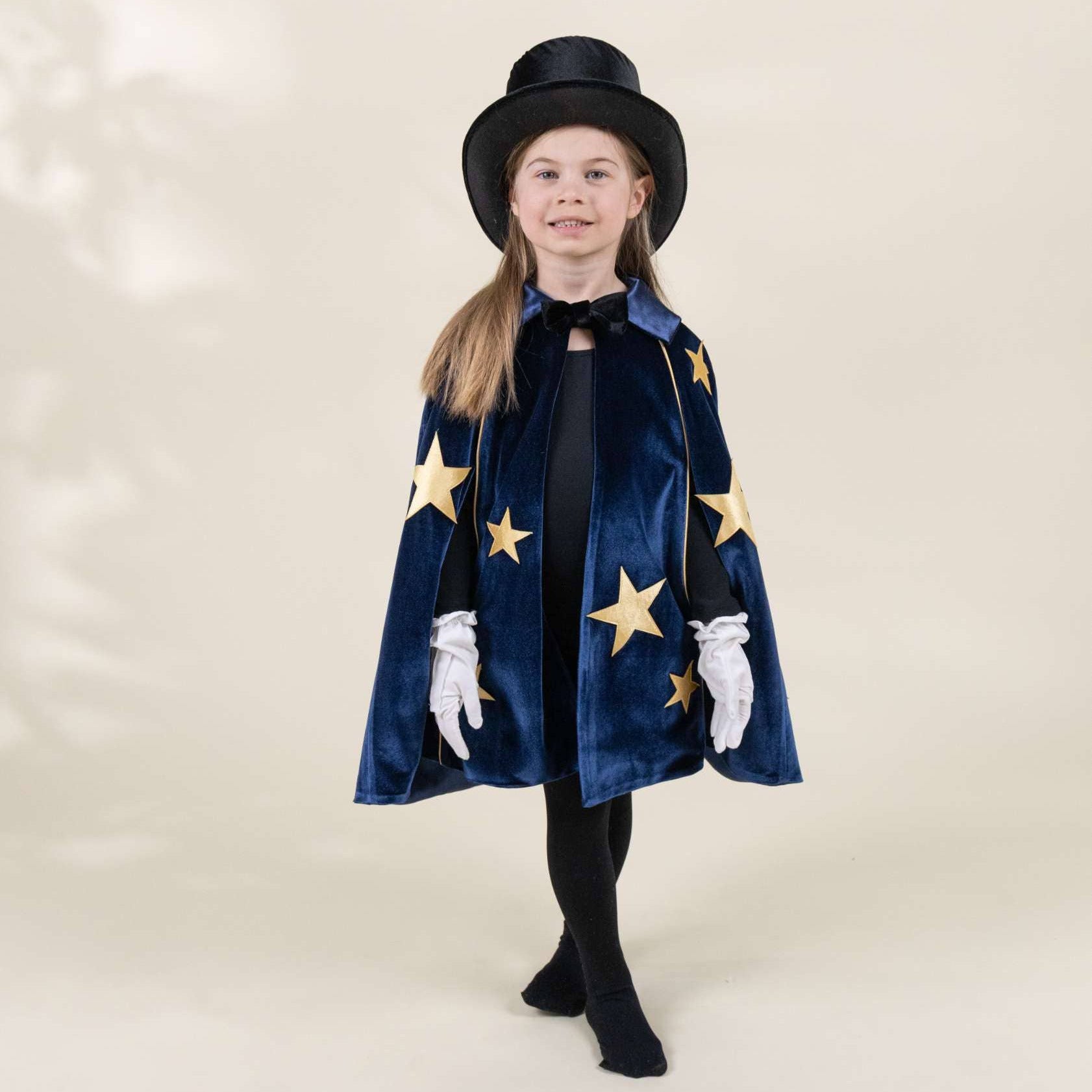 A young girl modeling a magician costume set, featuring a navy blue velvet cape with gold star appliqués, a matching black top hat, white gloves, and a black dress, set against a warm beige studio backdrop.