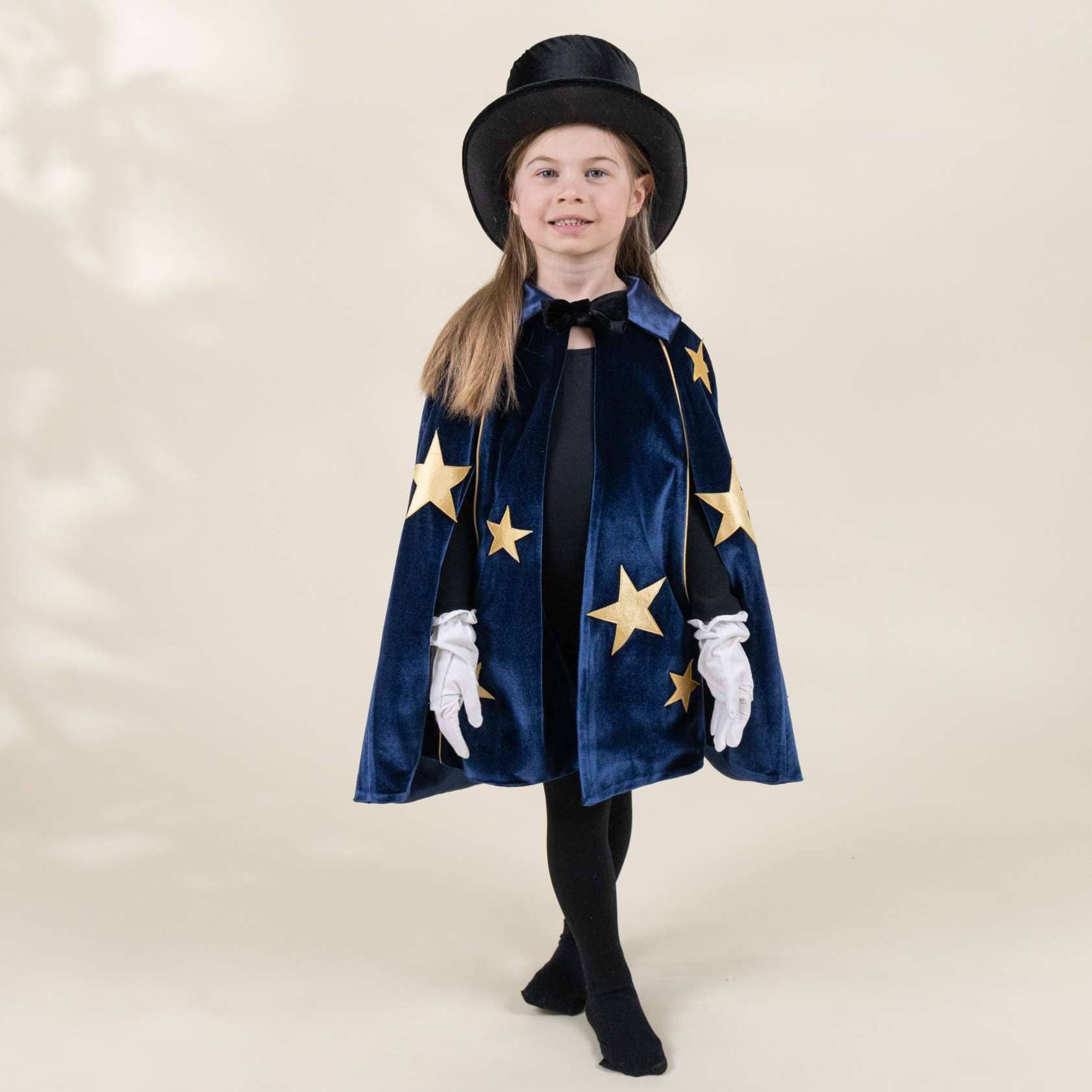 A young girl modeling a magician costume set, featuring a navy blue velvet cape with gold star appliqués, a matching black top hat, white gloves, and a black dress, set against a warm beige studio backdrop.