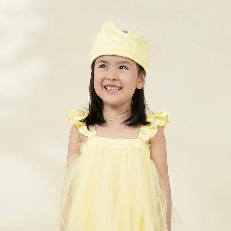 A young girl smiling and wearing a light yellow tulle dress with ruffled shoulder straps and a matching yellow bow headband, standing against a soft cream studio background.