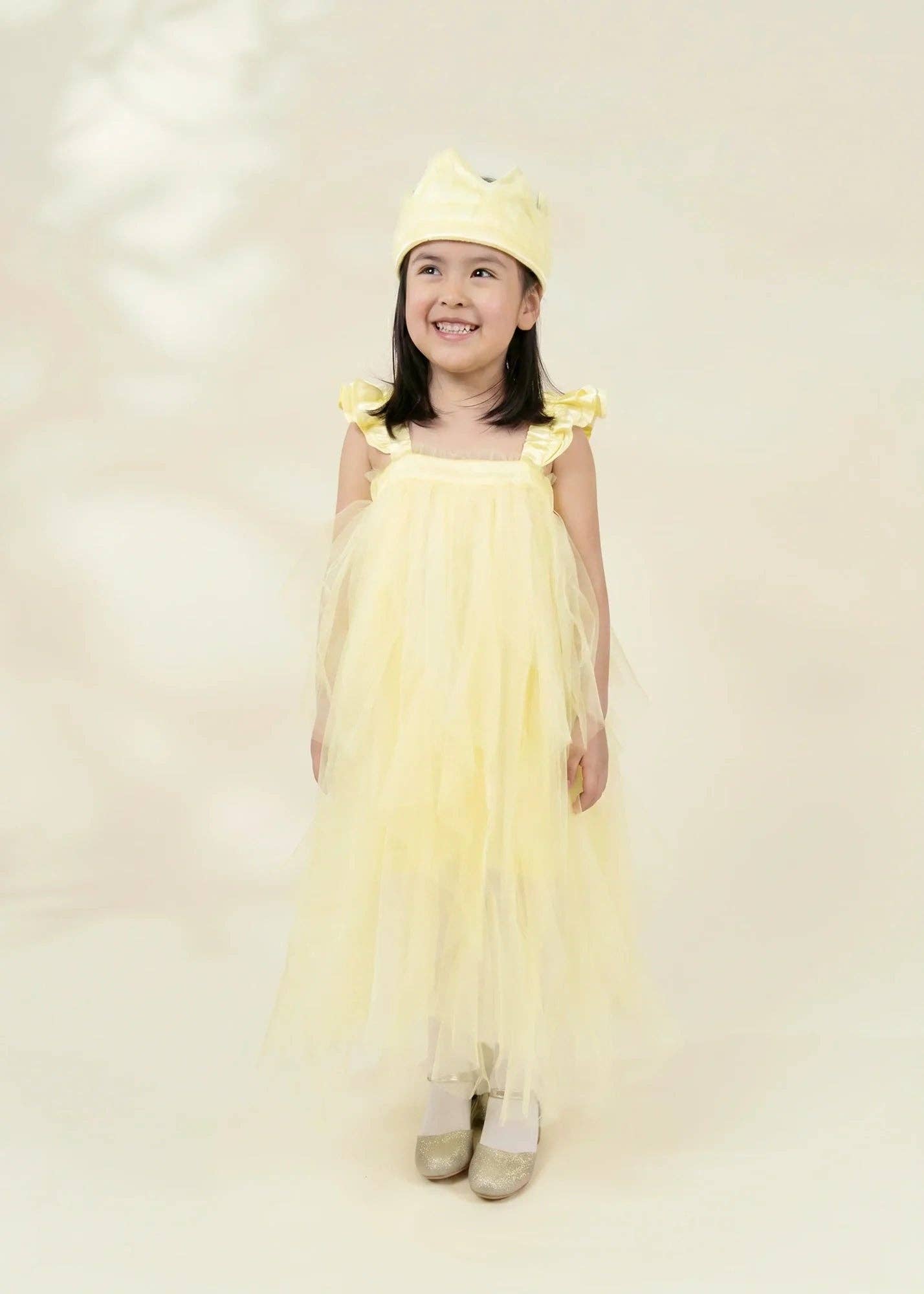 A young girl smiling and wearing a light yellow tulle dress with ruffled shoulder straps and a matching yellow bow headband, standing against a soft cream studio background.