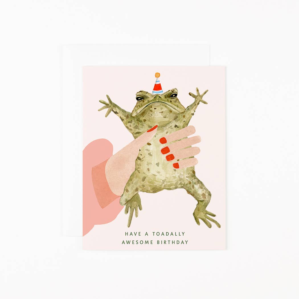 Illustration of a frog with a party hat held by a hand, with text 'Have a toadally awesome birthday' on a light pink background.