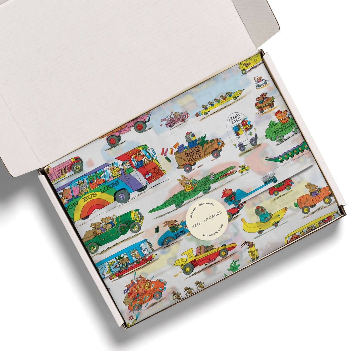 An open white cardboard gift box showing a colorful printed tissue with illustrated vintage buses, cars, crocodiles, and small characters on a pale background. A round sticker in the center reads 'RED CAP CLOTHING'.