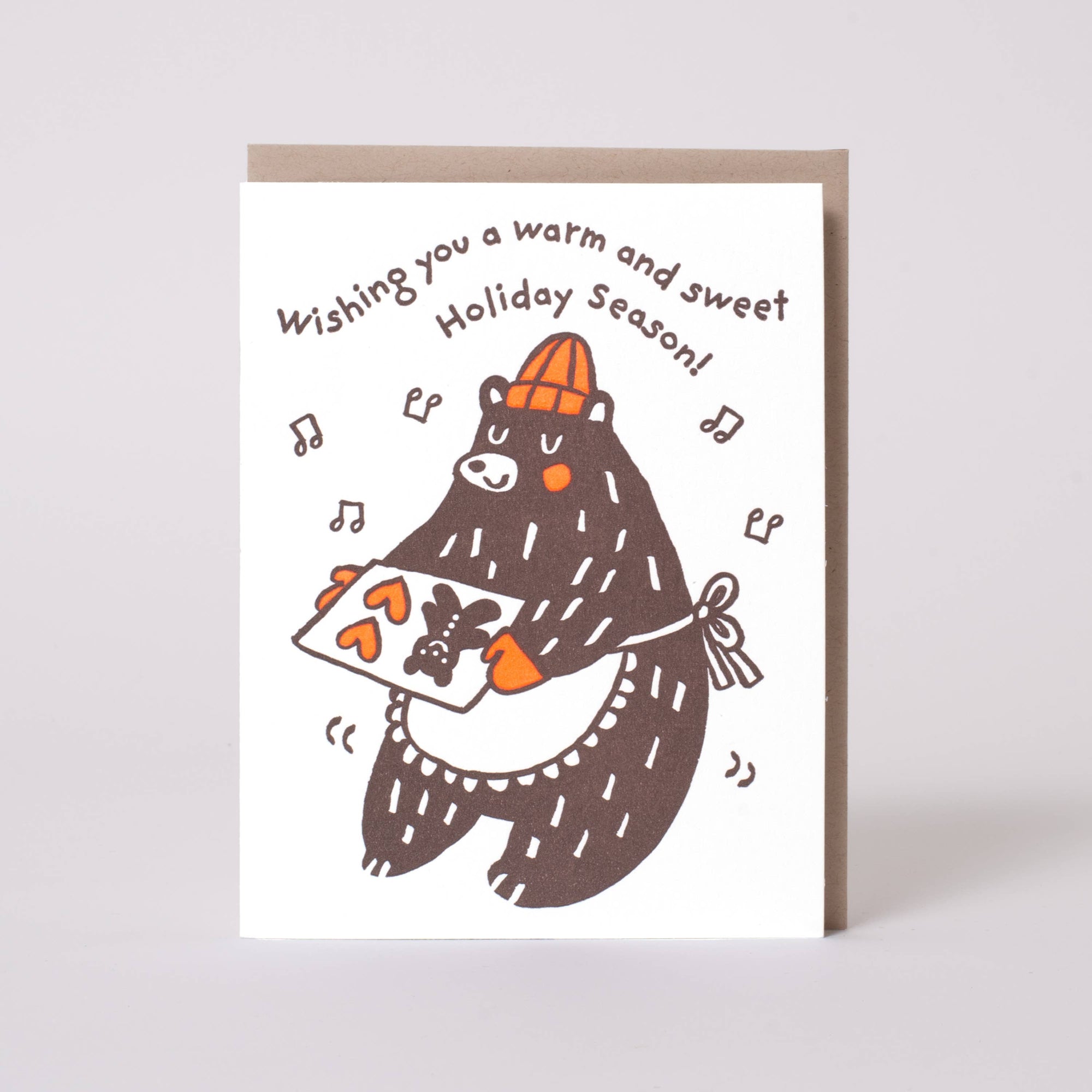 A white greeting card with a brown bear illustration holding a carrot, with the text 'Wishing you a warm and sweet Holiday Season!' above it. A brown envelope is partially visible behind the card against a light gray background.