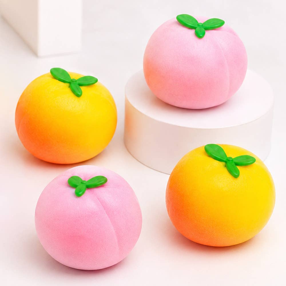 Four fruit-shaped squishy toys arranged on a white surface, including two pink peaches with green leaf tops and two yellow-orange citrus fruits with green leaf tops. One peach sits on a white cylindrical pedestal.