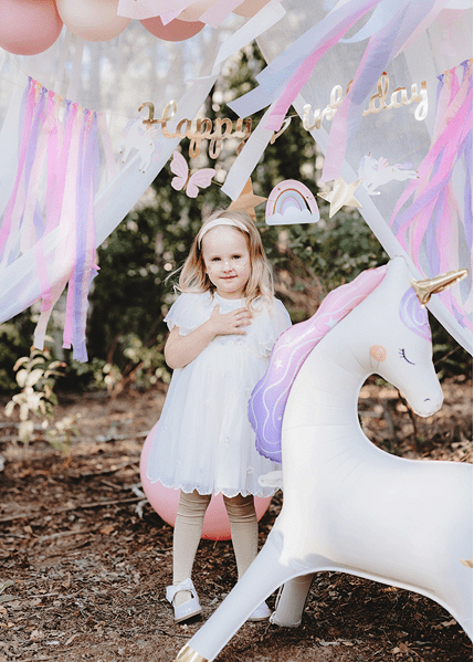 Young girl in a white dress standing next to a large inflatable unicorn under a 'Happy Birthday' sign with colorful decorations.