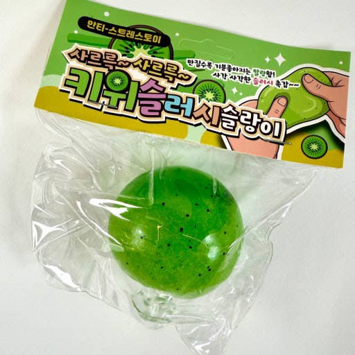 A green kiwi-shaped squishy toy sealed in clear plastic packaging with a green and yellow printed header card featuring Korean text and kiwi illustrations.