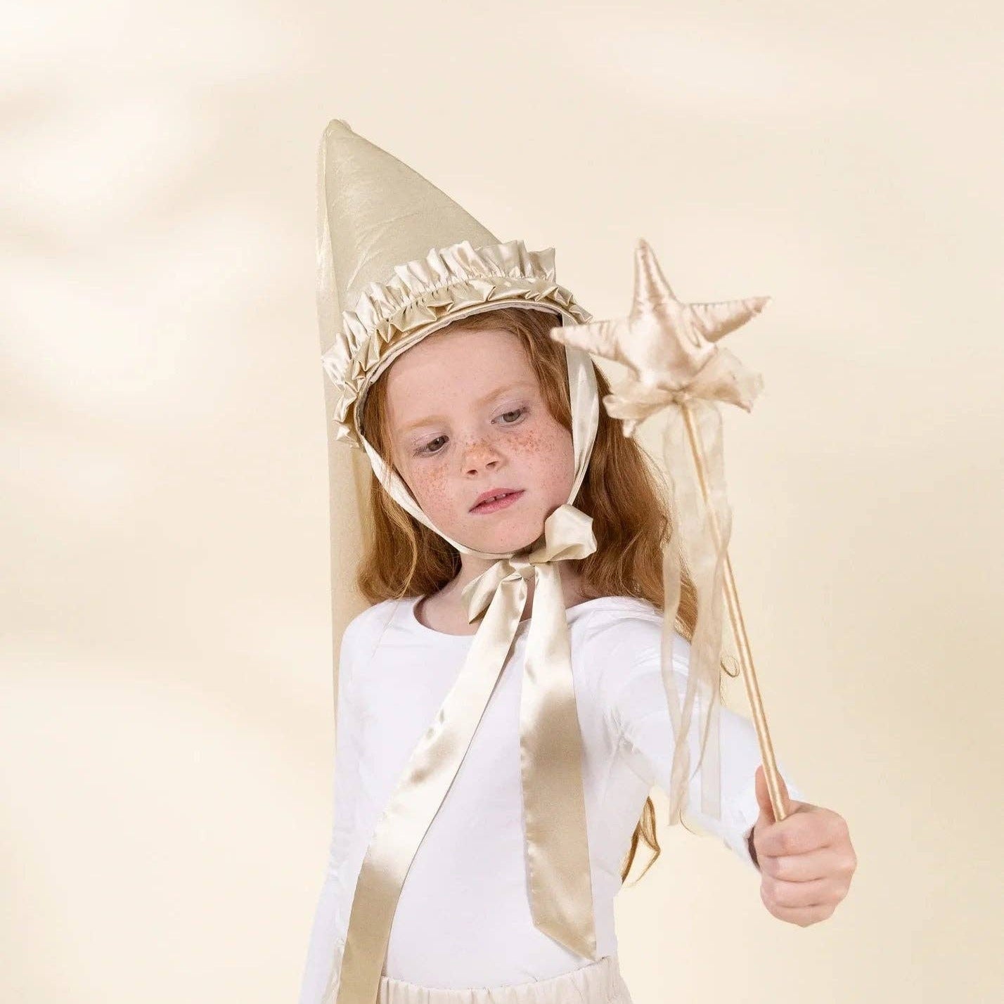 A young girl wearing a gold and white wizard costume, featuring a pointed gold hat with a white ruffled trim, a matching satin ribbon, and holding a gold star wand, set against a plain beige studio background.