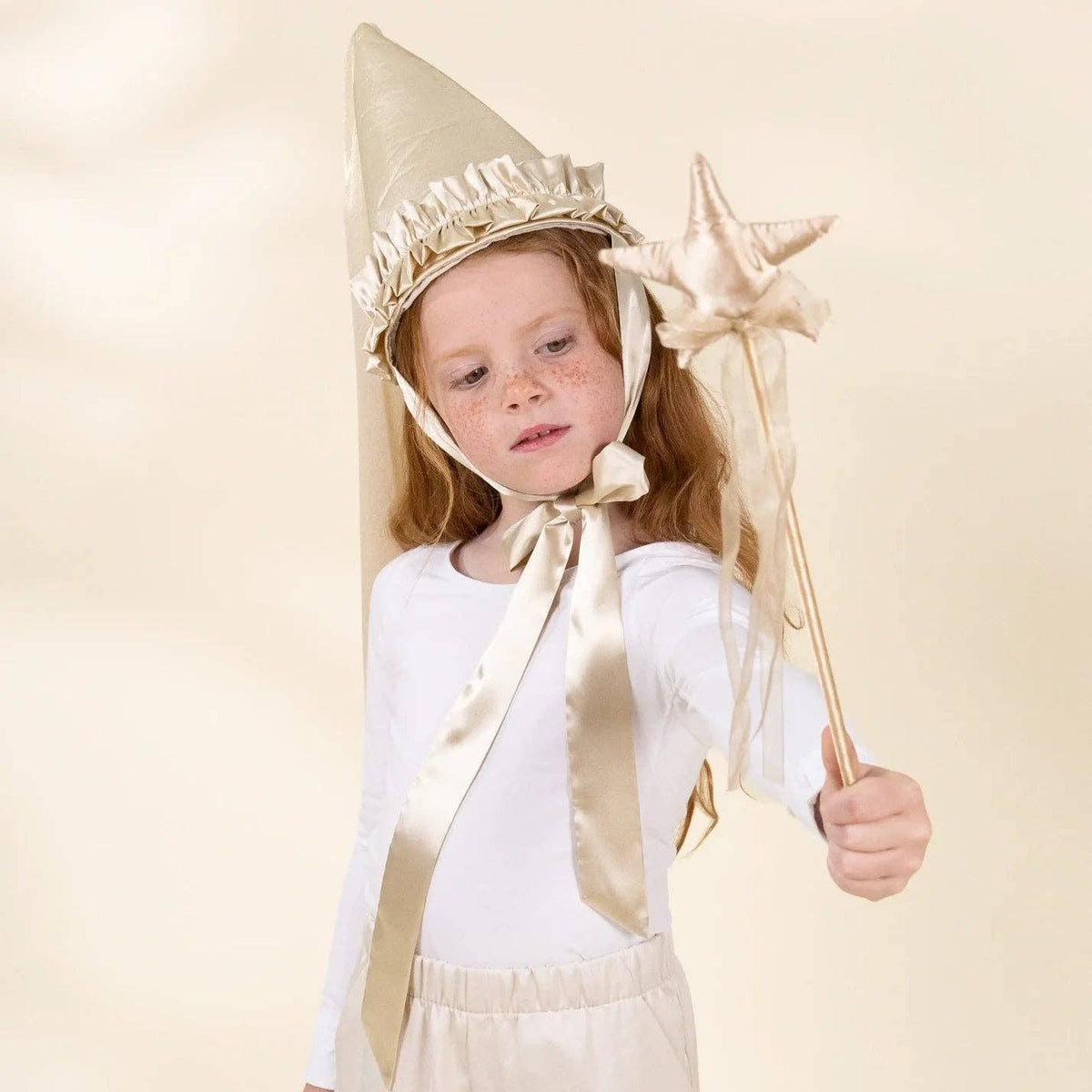 A young girl wearing a gold and white wizard costume, featuring a pointed gold hat with a white ruffled trim, a matching satin ribbon, and holding a gold star wand, set against a plain beige studio background.