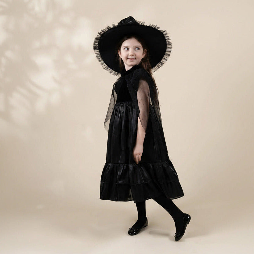 A young girl modeling a black dress with a wide-brimmed black hat featuring white ruffled trim, standing against a solid beige background.
