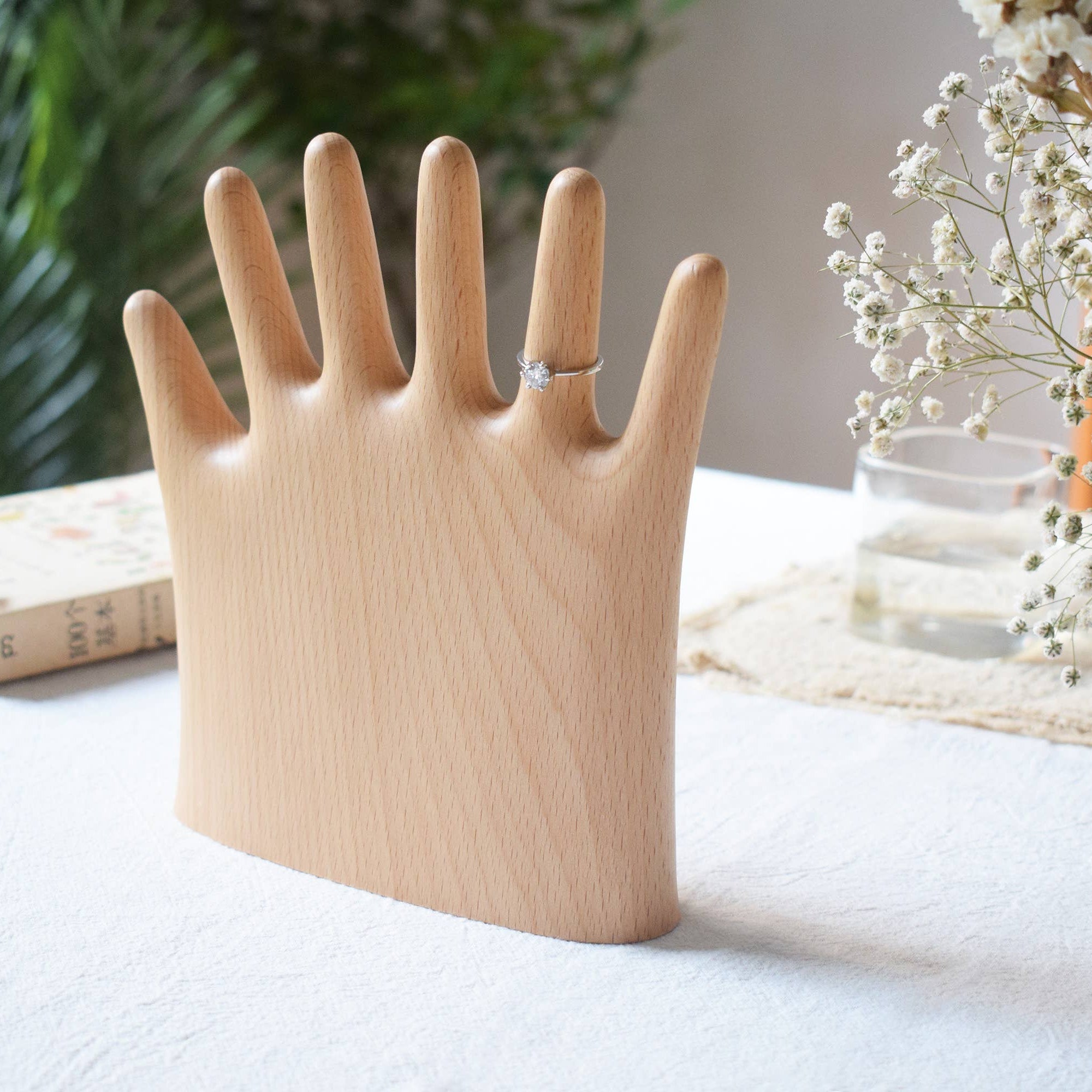 Wooden ring holder shaped like a hand on a table with flowers and books in the background