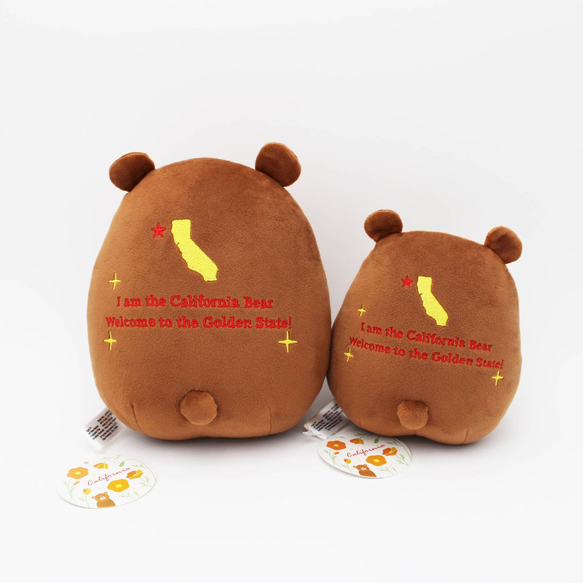 Two brown plush bear-shaped pillows shown from the back on a white background, one larger and one smaller, both printed with a yellow California map outline, red text reading 'I am the California Bear Welcome to the Golden State!', and small yellow star accents. A small round tag is visible in front of each pillow.