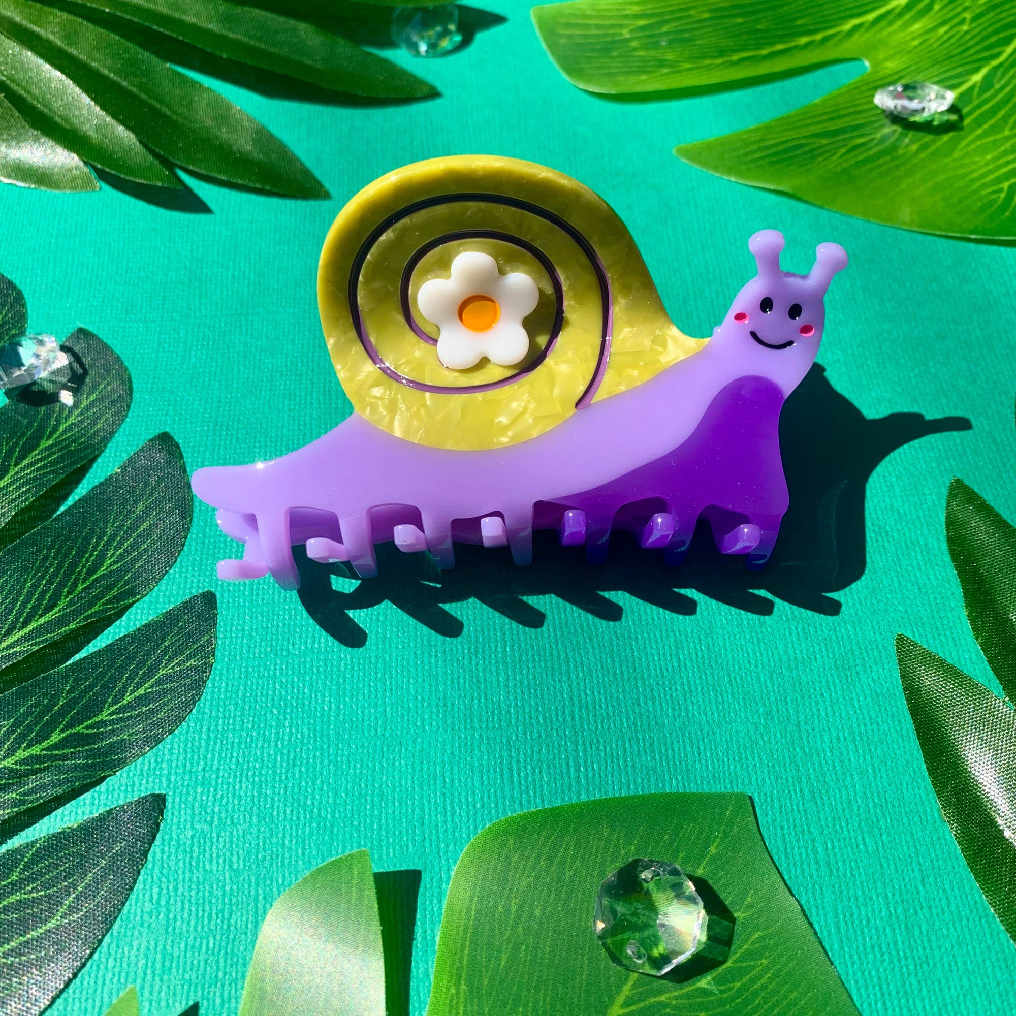 Purple snail-shaped hair clip with a yellow shell and white flower on a green leaf background