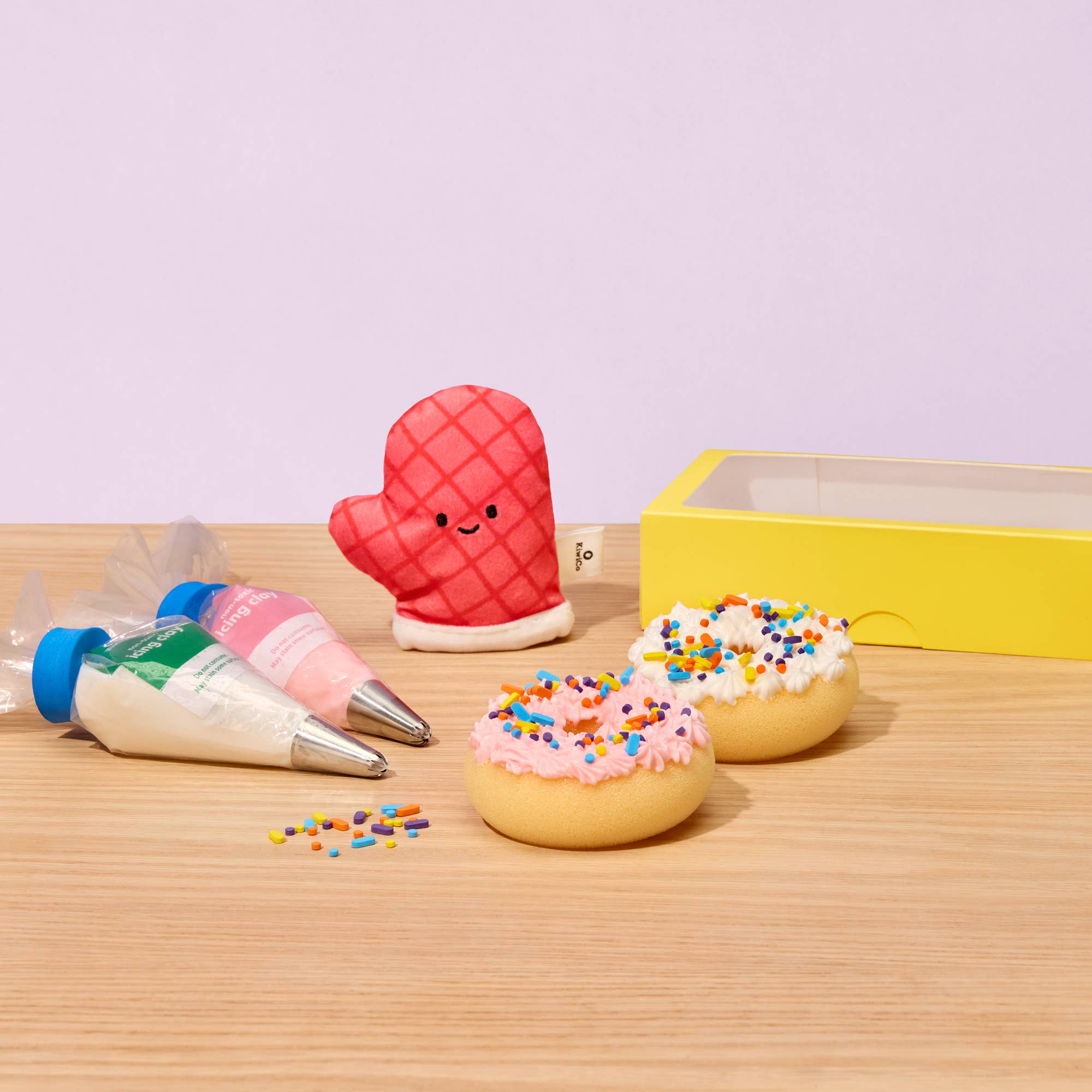 A cute kitchen-themed toy set on a light wood table against a lavender background, featuring a red quilted oven mitt character, two donut-shaped toys with pink and white frosting and multicolor sprinkles, two piping bags filled with icing, and a yellow gift box.