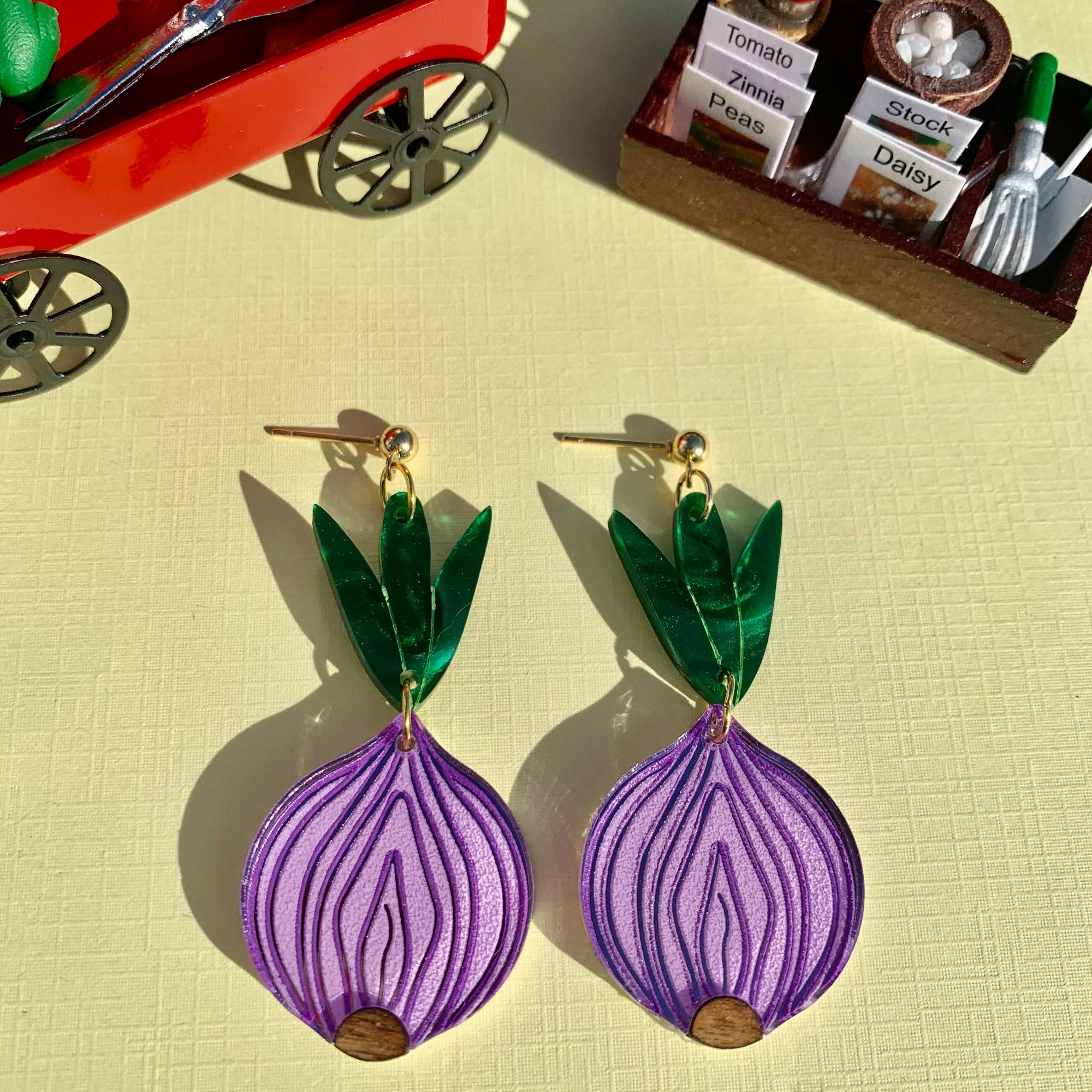 Purple onion-shaped earrings with green leaves on a yellow surface, with a toy garden set in the background.