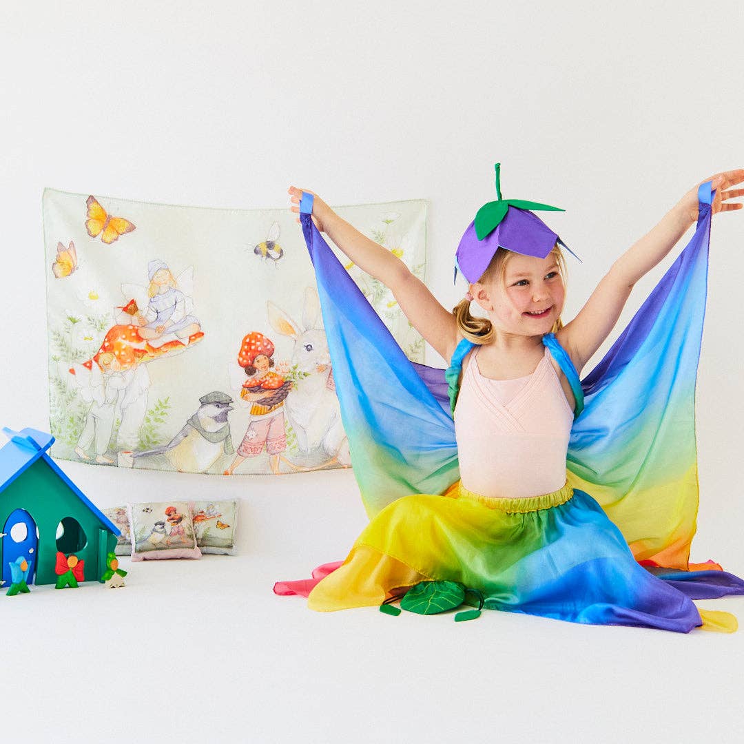 Child in a colorful costume with a rainbow scarf in a room with children's artwork on the wall.