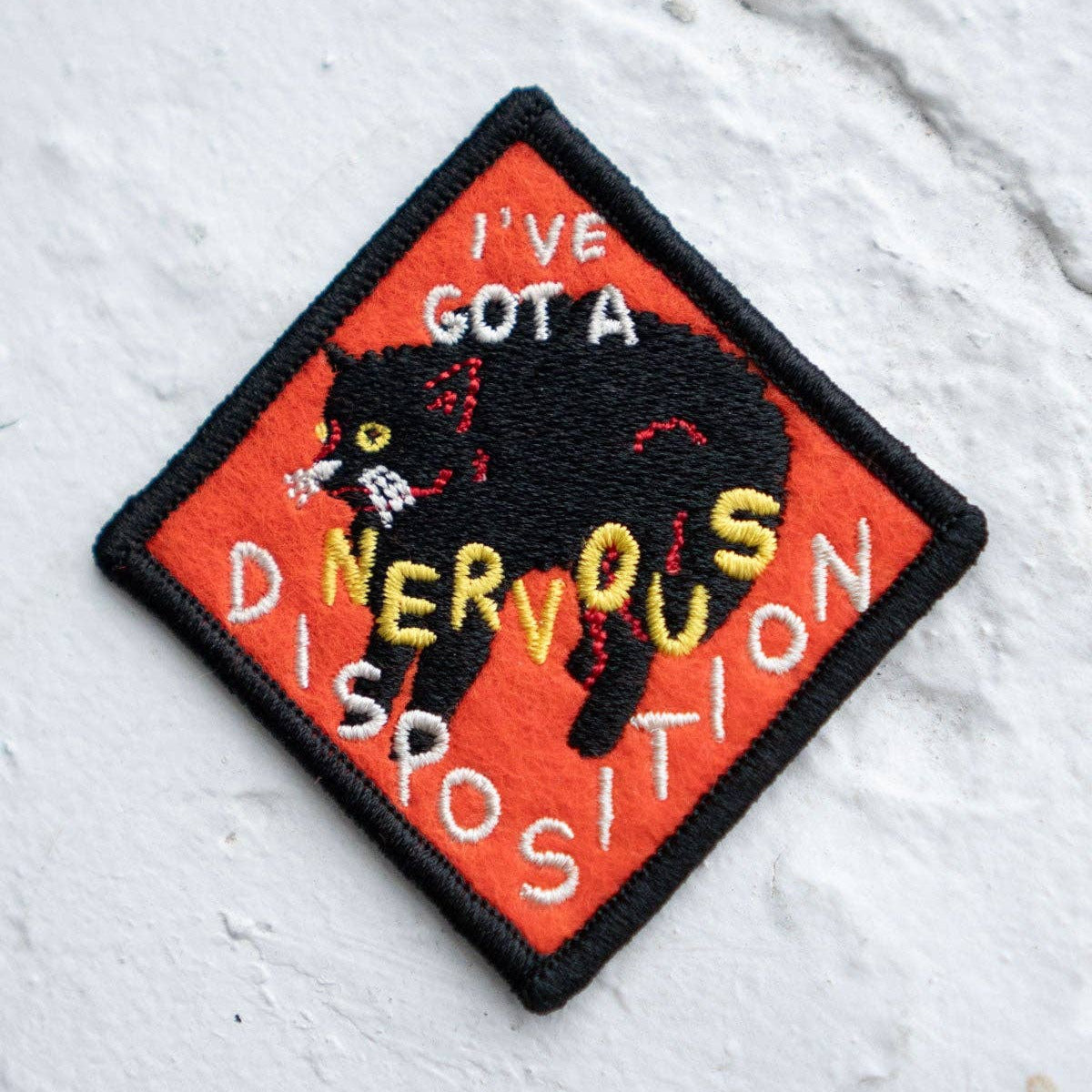 Embroidered patch with a black cat and text on an orange background, placed on a textured white surface.