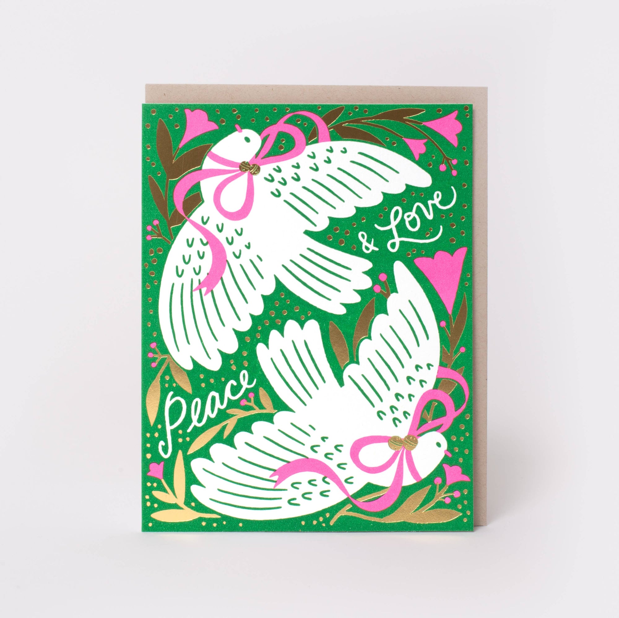 A greeting card with a green background featuring two white doves wearing pink bows, with the words 'Peace & Love' in white script, shown with a kraft envelope behind it on a light gray background.