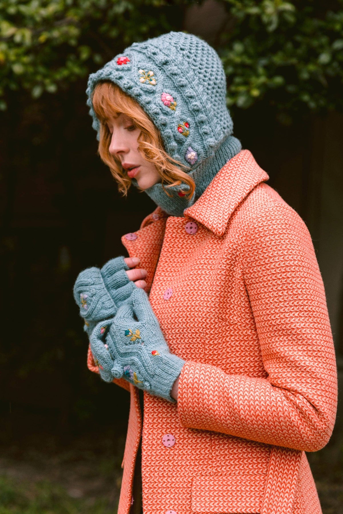 A woman wearing a textured coral-orange coat, a dusty blue crocheted bonnet with colorful embroidered flowers, and matching blue knit mittens, standing outdoors against a dark, blurred background.