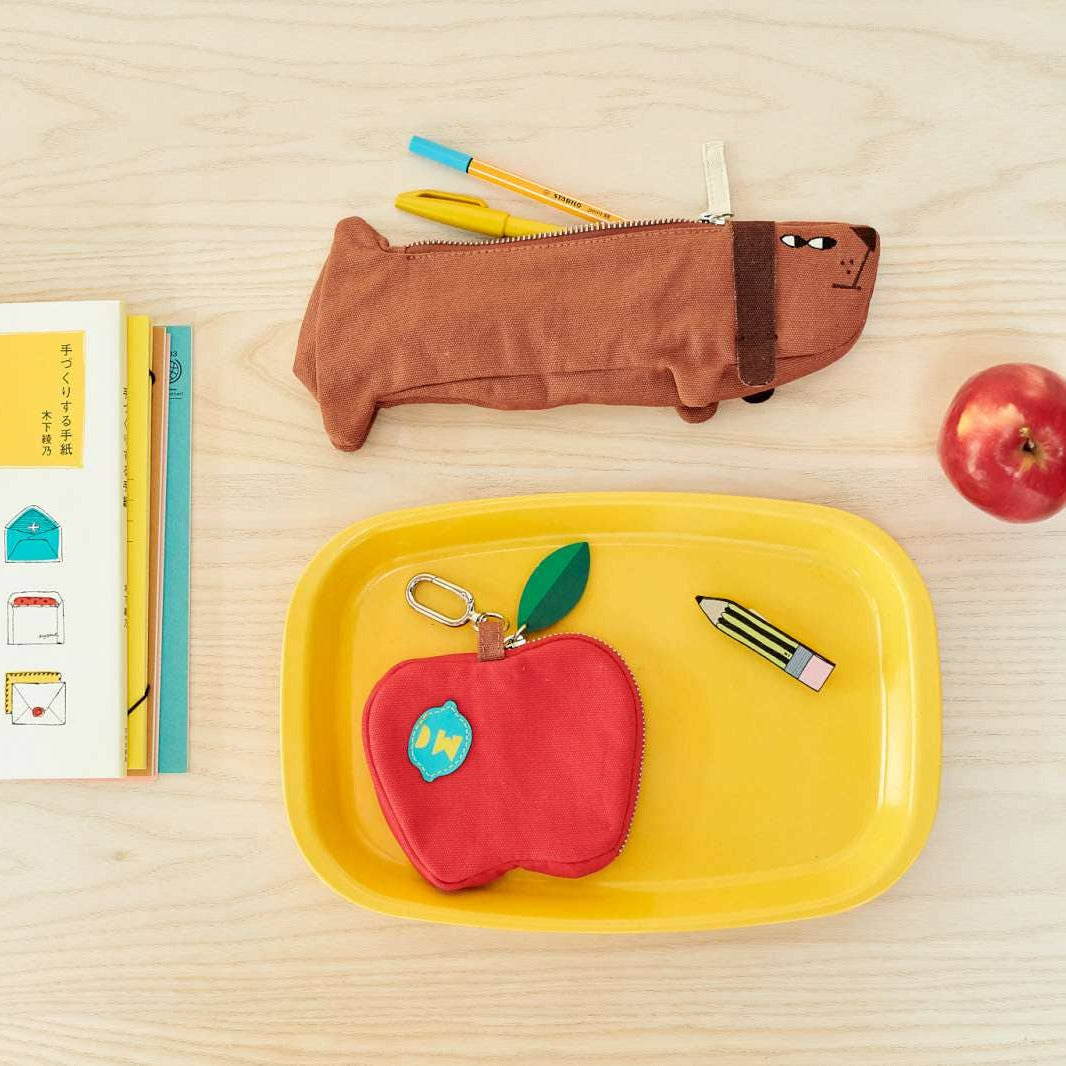 A styled flat-lay of school or office accessories on a light wood surface: a brown dachshund-shaped pencil case, a red apple-shaped keychain pouch on a yellow tray, a white notebook with colorful envelope illustrations, and a red apple.