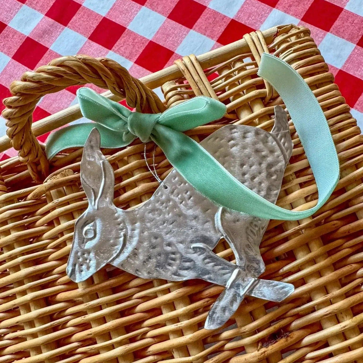 Decorative metal hares with a green ribbon on a wicker basket against a red and white checkered background.