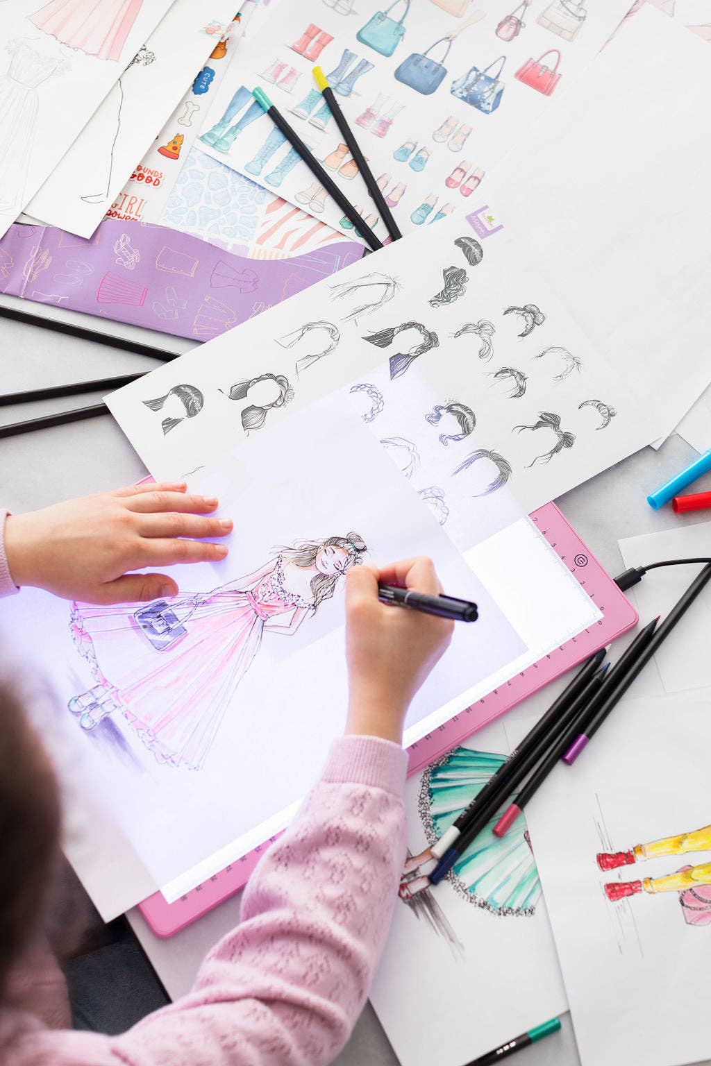 Child drawing fashion illustrations with markers on a table.
