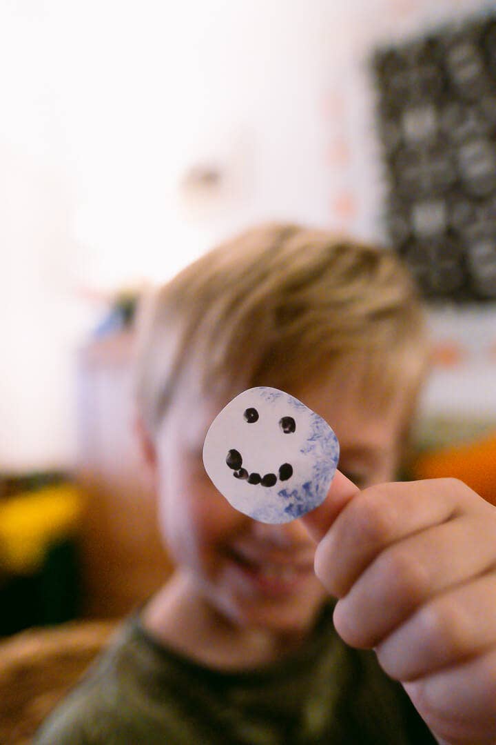 A child holds up a small round white craft piece with black painted facial features, with the child's face blurred in the background.