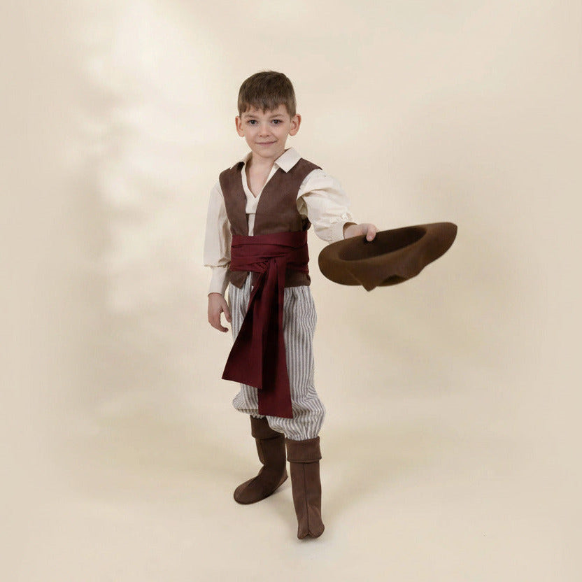 A young boy modeling a pirate costume consisting of a brown vest, white long-sleeve shirt, striped pants, a dark red sash, and brown boots, while holding a brown hat against a solid beige background.